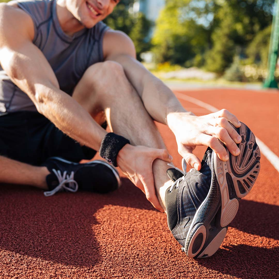 Um homem com roupa desportiva sentado numa pista de corrida, agarrado ao tornozelo esquerdo com as duas mãos. Parece estar a alongar ou com cãibras. Está a usar uma t-shirt cinza sem mangas e ténis de corrida pretos. 